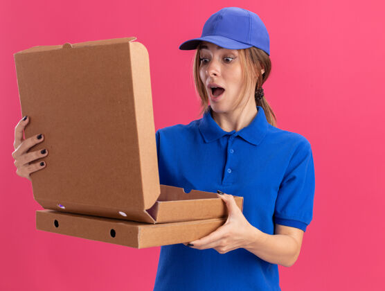 表情穿着制服的年轻漂亮的送货员兴奋地抱着披萨盒 看着隔离在粉红色墙上的披萨盒兴奋人披萨