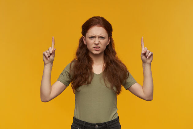 面部消极 皱眉的女人 长着姜黄色的头发穿着绿色t恤人和情感的概念指着复制空间 隔离在橙色的墙上年轻消极表情