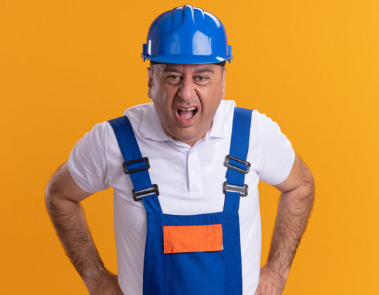 脸恼怒的成年建筑工人穿着制服看着橙色墙壁上孤立的正面成年人制服建设者