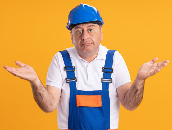 脸困惑的成年建筑工人穿着制服 双手张开孤立地站在橙色的墙上感情困惑姿势