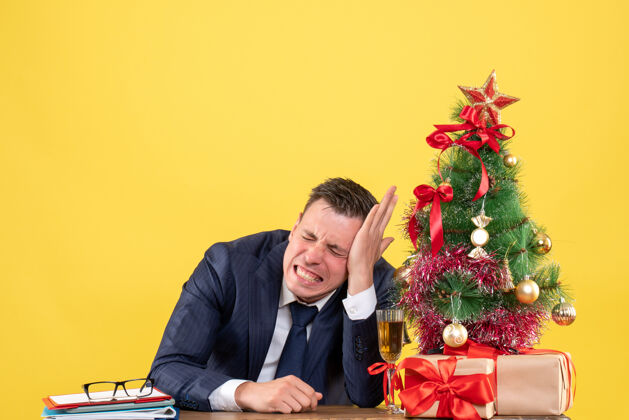 男人前视图焦躁不安的男子闭着眼睛坐在圣诞树旁的桌子上 黄色背景上有礼物前面生意关门