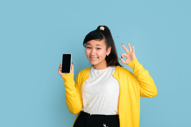 亚洲人在蓝色工作室背景上孤立的亚洲青少年肖像美丽的深色长发女性模特人类情感的概念 面部表情 销售 广告显示空白的手机屏幕人表情帅气