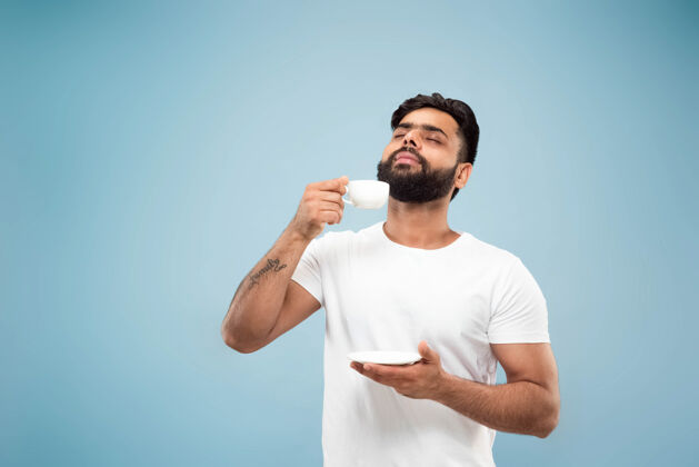 成功蓝色背景上穿着白衬衫的印度年轻人的半身特写肖像人类情感 面部表情 销售 广告概念负面空间喝咖啡或茶的享受人肖像选择