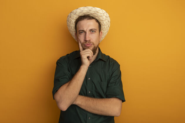 人困惑的金发帅哥戴着沙滩帽把手放在橘色墙上孤立的下巴上帅哥男人站