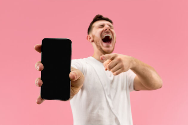 脸年轻帅哥在粉色背景上展示智能手机屏幕 一张令人惊讶的脸人类情感 面部表情概念时尚色彩手机惊讶屏幕
