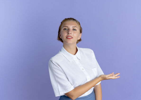 手年轻自信的俄罗斯金发女孩用手指着侧面 看着隔离在紫色背景上的相机 还有复制空间金发俄罗斯相机