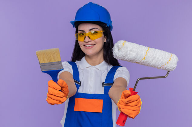 手套年轻的女建筑工人穿着施工制服 戴着安全帽 戴着橡胶手套 拿着油漆辊和刷子头盔站立刷子