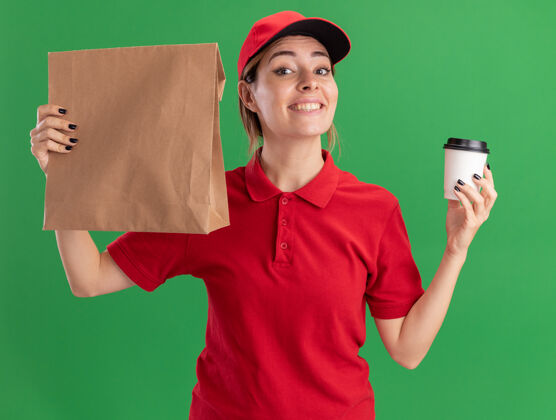 递送微笑着的年轻漂亮的女送货员穿着制服 把纸杯和纸包装隔离在绿色的墙上微笑抱着包装