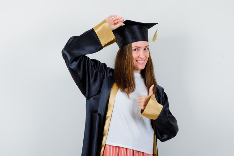 头女毕业生竖起大拇指 手放在头上 穿着制服 穿着休闲服 看上去很开心 正面照帽微笑成功