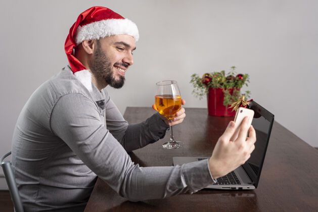 杯子特写镜头一个西班牙裔男子戴着圣诞老人的帽子 享受他的酒和自拍男人圣诞老人桌子