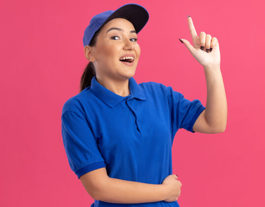 帽子身穿蓝色制服 戴着帽子的年轻女送货员站在粉色的墙上 开心而积极地看着前面 露出食指有了新的想法显示制服交货