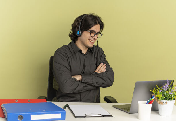 办公室戴着耳机 戴着眼镜 面带微笑的年轻上班族坐在办公桌旁 手里拿着办公工具 双手交叉看着笔记本电脑 背景是绿色的 还有复印空间光学微笑耳机