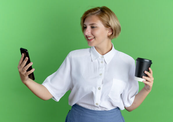 女孩年轻的微笑的金发俄罗斯女孩拿着咖啡杯看着手机在绿色的背景与复制空间隔离俄罗斯人微笑电话