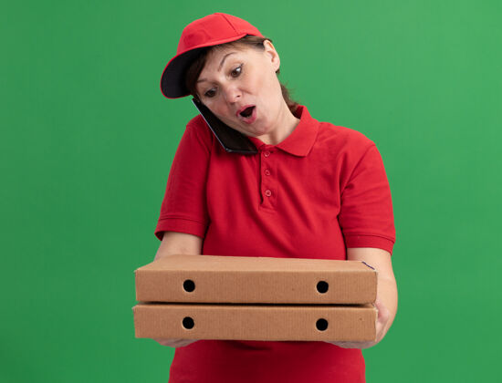 困惑身穿红色制服 戴着帽子 拿着披萨盒的中年女送货员站在绿色的墙上 神情迷茫地用手机交谈拿着送货帽子
