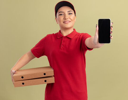 欢呼身穿红色制服 头戴鸭舌帽 拿着披萨盒 拿着手机的年轻送货员站在绿色的墙上 面带微笑地看着前方手机送货年轻人