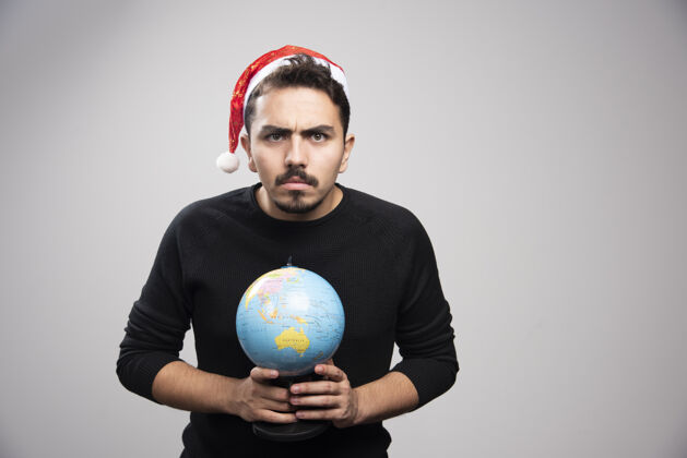 站立戴着圣诞老人帽子拿着地球仪的愤怒男人圣诞时尚肖像