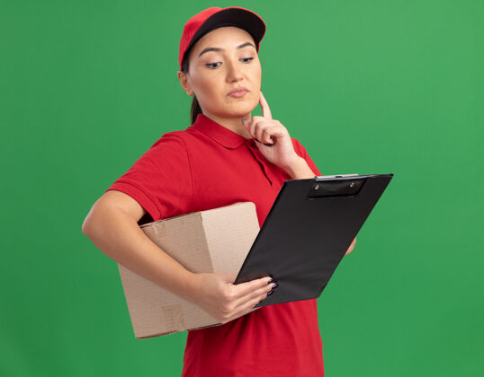 年轻身穿红色制服 头戴帽子的年轻女送货员拿着一个带剪贴板的纸板箱 带着沉思的表情站在绿色的墙上看着它立场女人盒子