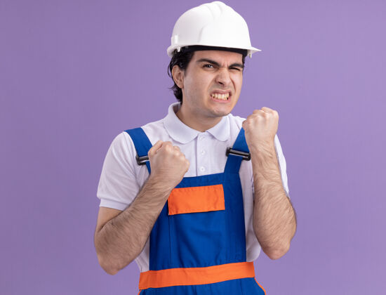 握紧愤怒的年轻建筑工人身穿建筑制服 戴着安全帽 握紧拳头 站在紫色的墙上 既恼火又恼火恼怒年轻愤怒