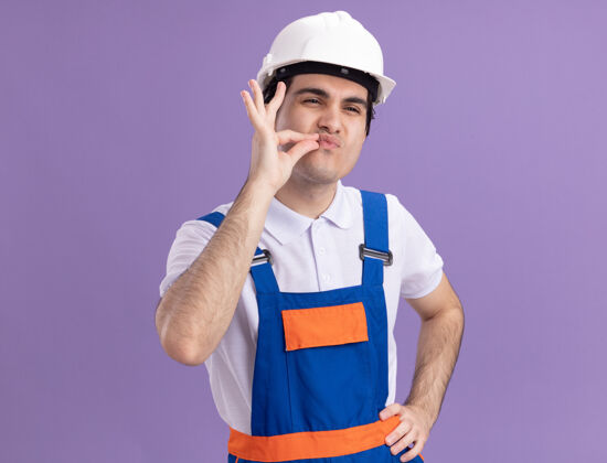 手势身穿施工制服 头戴安全帽的年轻建筑工人用手指做着沉默的手势 就像在紫色的墙上拉着拉链闭上了嘴站立手指建筑