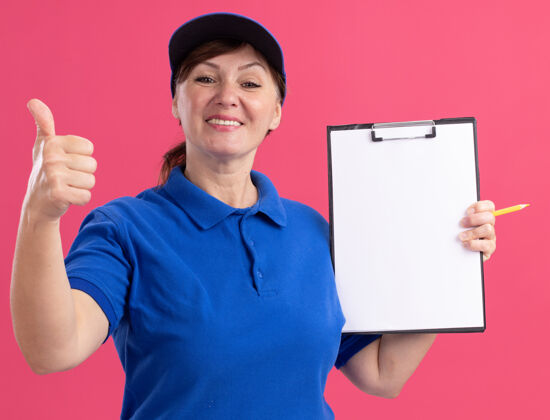 中间中年女送货员身穿蓝色制服 戴着帽子 手里拿着写字板 手里拿着空白页 面带微笑地站在粉红色的墙上竖起大拇指拇指剪贴板交货