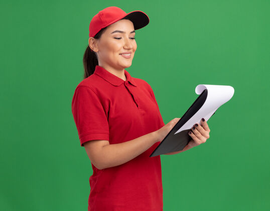 剪贴板身着红色制服 头戴便笺帽 脸上挂着微笑 站在绿色墙壁上的快乐的年轻女送货员交货微笑帽子