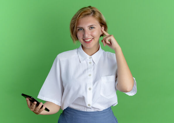 电话年轻的金发碧眼的俄罗斯女孩拿着电话 把手指放在耳朵上 绿色的背景上 留有复制空间手指微笑耳朵