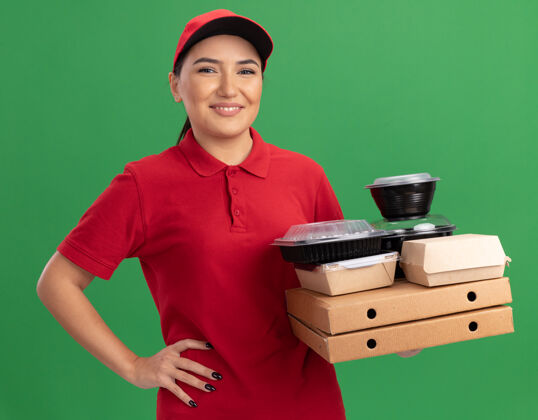 盒子身穿红色制服 头戴鸭舌帽的年轻女送货员拿着比萨饼盒和食品包 站在绿色的墙上 面带微笑地看着前方送货抱着站着