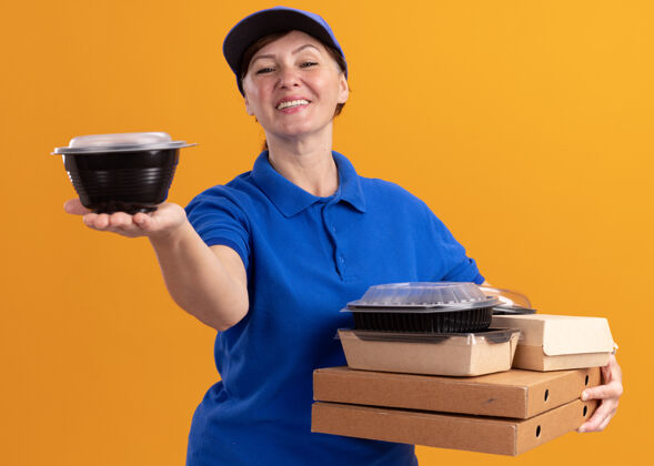 制服身穿蓝色制服 头戴鸭舌帽的中年女送货员拿着披萨盒和食品包 微笑着自信地站在橙色的墙上看着前方微笑站着盒子