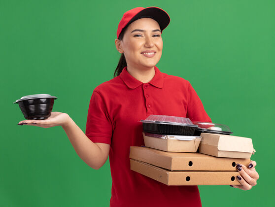 欢呼身穿红色制服 头戴鸭舌帽的年轻女送货员拿着比萨饼盒和食品包 站在绿色的墙上 面带微笑地看着前方送货制服帽子