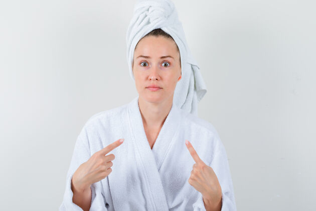 乐趣年轻女子指着自己在白色浴衣 毛巾和困惑的神情 前视图床她自己时尚