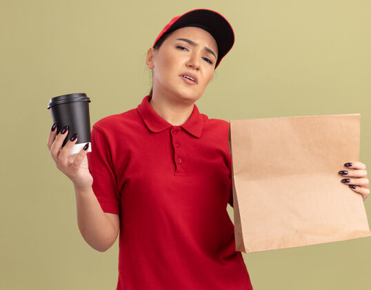 站着年轻的送货员身穿红色制服 戴着帽子 手里拿着纸包和咖啡杯 站在绿色的墙上 不高兴地看着前面拿着送货帽子