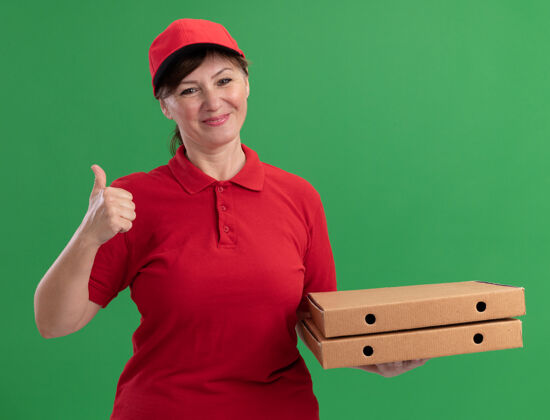 盒子快乐的中年送货员 身穿红色制服 戴着帽子 拿着披萨盒 面带微笑 站在绿色的墙上竖起大拇指表演快乐拇指