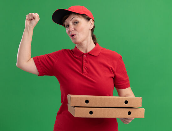 帽子身穿红色制服 头戴鸭舌帽的中年女送货员举着披萨盒望着前面举起的拳头幸福而自信地站在绿色的墙上中年拳头信心