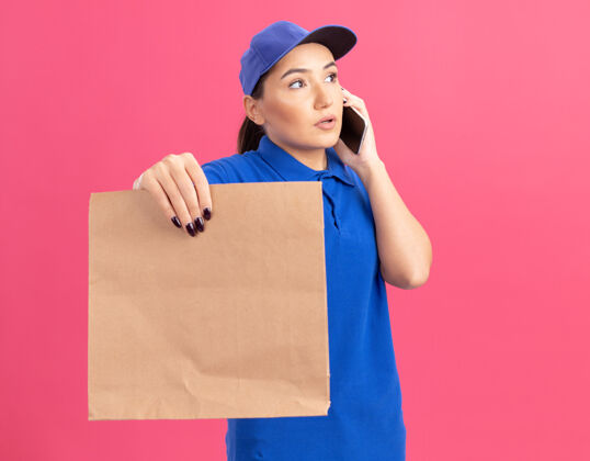 女人站在粉红色的墙上 一个身穿蓝色制服 戴着帽子 拿着纸包 拿着手机的年轻送货员看起来很困惑年轻帽子包装