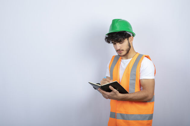 工作帅气的男工程师戴着安全帽在白墙上的笔记本上写字高质量的照片制服钢笔帽子