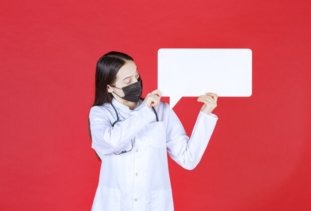 从业者女医生带着听诊器 戴着黑色面罩 手里拿着一张长方形的问讯台制服疾病董事会