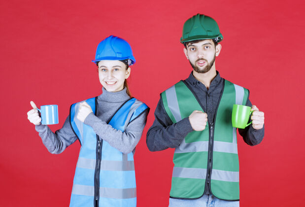 年轻人男女工程师戴头盔 手持蓝绿色马克杯 并显示满意标志休闲年轻咖啡