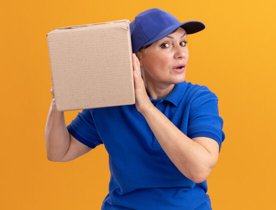 纸板身穿蓝色制服 头戴鸭舌帽的中年女送货员站在橙色的墙上 用硬纸盒捂着耳朵听着盒子送货女人