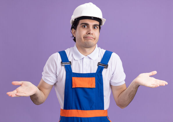 头盔身穿建筑制服 戴着安全帽的年轻建筑工人站在紫色的墙上 困惑地向两边伸出双臂 没有回答伸展安全侧线