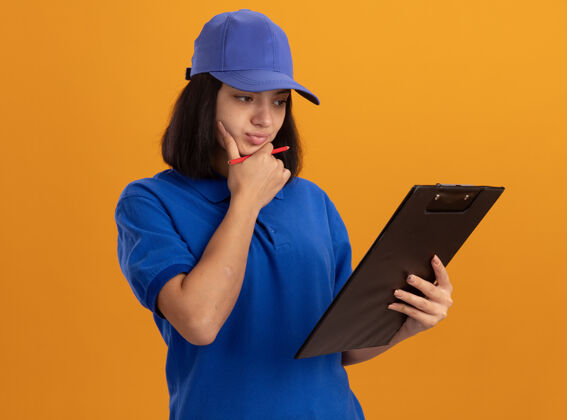 女孩身穿蓝色制服 戴着帽子的年轻送货员站在橙色的墙上 带着沉思的表情看着它帽子剪贴板年轻