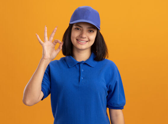 帽子身着蓝色制服 戴着帽子的年轻送货女孩站在橙色的墙上 脸上挂着“ok”的牌子女孩制服接生