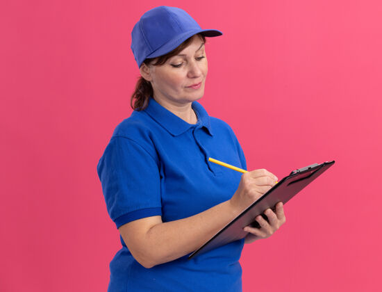 制服身穿蓝色制服 戴着帽子 拿着剪贴板和铅笔的中年女送货员站在粉红色的墙上 表情严肃女人站着严肃