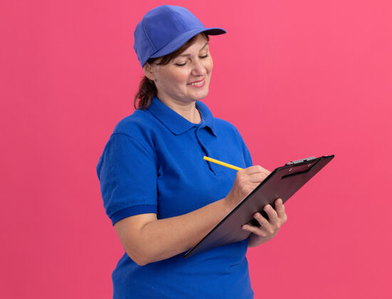 铅笔身穿蓝色制服 头戴鸭舌帽的中年送货妇女站在粉红色的墙上 手里拿着剪贴板和铅笔 脸上带着微笑帽子举行微笑
