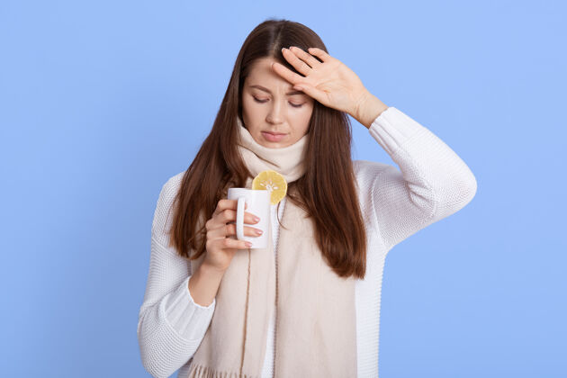 围巾流感治疗画像中生病的年轻女子裹着围巾喝着热茶饮料 得了季节性流感 病毒感染 摆出隔离在蓝色墙上的姿势治疗药丸额头