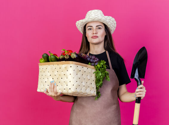 制服看着镜头美丽的园丁女孩穿着制服 戴着园艺帽 拿着蔬菜筐 铁锹孤立在粉红色的背景上黑桃花园相机