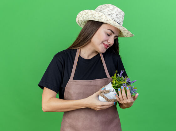 花园高兴美丽的园丁女孩穿着制服 戴着园艺帽 手持花盆 看着隔离在绿色背景上的花朵手持请戴制服