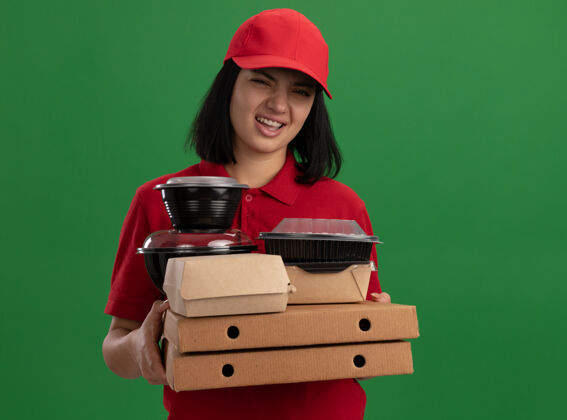 食物身穿红色制服 头戴鸭舌帽 手持披萨盒和食品包的年轻送货女孩站在绿色的墙上 脸上带着失望的表情拿着盒子女孩