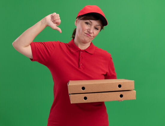 中间身穿红色制服 头戴鸭舌帽的中年女送货员拿着比萨饼盒望着前面 微笑着指着站在绿色墙壁上的自己盒子制服女人