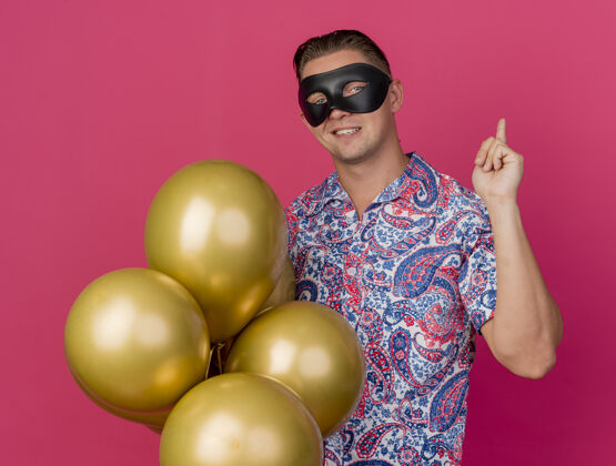 戴着高兴的年轻人戴着化装眼罩拿着气球 点在后面孤立的粉红色背景面具眼睛年轻
