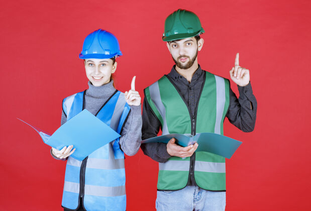 工人戴着头盔的男女工程师拿着蓝色文件夹 读着它 发着通知问题年轻人检查表